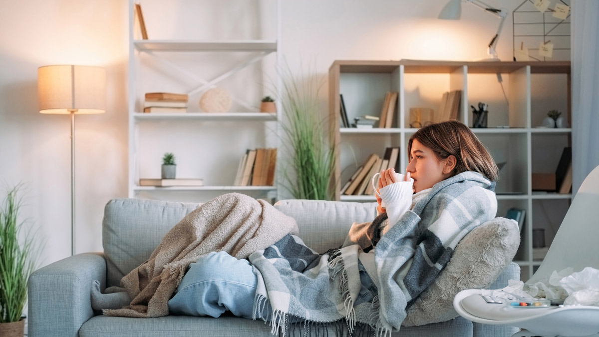 Woman reclines on a couch, bundled in a blanket, sipping tea from a mug