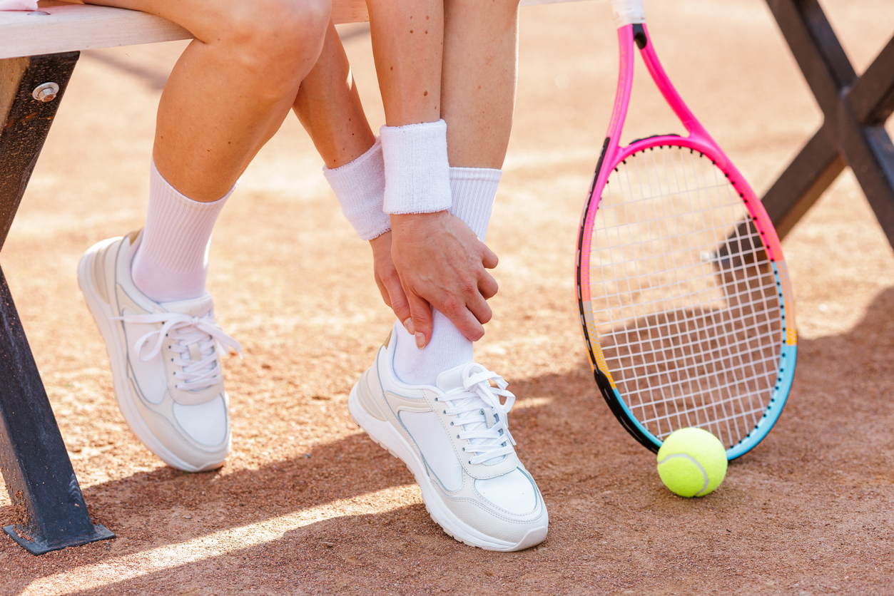 Tennis player holding sore ankle while sitting on bench with racket nearby